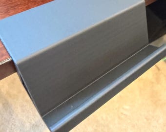 Op de afbeelding: Een matgrijs, L-vormig bureauaccessoire met een plat bovenoppervlak en een opstaande voorrand die is ontworpen om over de rand van een tafel te haken. Het object heeft een gladde, solide constructie met een strak, hoekig profiel.