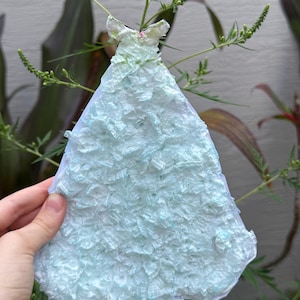 Puede incluir: Un vestido de papel azul claro con falda y corpiño con volantes. El vestido está sostenido por una mano y se encuentra sobre un fondo de plantas verdes y una pared blanca. El vestido es un proyecto de manualidades.