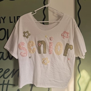 May include: A white short-sleeved t-shirt featuring the word senior appliqued in pastel-colored, patterned fabric letters. The design is accented with small fabric flowers, each centered with a button, and scattered embroidered star details.