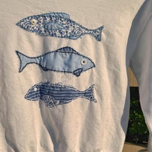 May include: A white long-sleeve sweatshirt featuring three appliquéd fish designs arranged vertically. Each fish is crafted from different blue fabric patterns, including floral, solid light blue, and striped, with small white buttons for eyes.