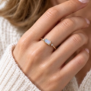 Könnte beinhalten: Ein Goldring mit einem rechteckigen Opal-Mittelstein mit Perlenrand, flankiert von zwei kleinen runden, klaren Steinen auf einem dünnen Goldband. Der Ring wird an einem Finger getragen und zeigt die schillernden Farben des Opals.