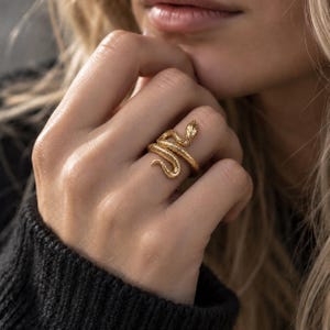 Könnte beinhalten: Ein goldfarbener Ring in Form einer gewundenen Schlange mit einer strukturierten Oberfläche, die Schuppen nachahmt. Der Schlangenkopf befindet sich oben und der Körper windet sich um den Finger, wodurch ein durchgehendes, geschwungenes Band entsteht.