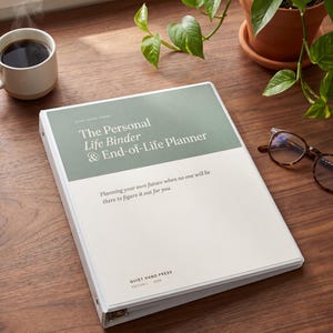 Puede incluir: Un archivador blanco titulado "The Personal Life Binder & End-of-Life Planner" está sobre una superficie de madera. El archivador tiene una cubierta verde y blanca con texto. Una taza de café, gafas y una planta en maceta también están presentes.