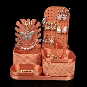 May include: A copper-colored jewelry organizer featuring a leaf-shaped necklace stand, a rectangular pegboard for earrings, and two storage bins with textured sides. One bin contains multiple ring cones, while the other holds a pearl bracelet.