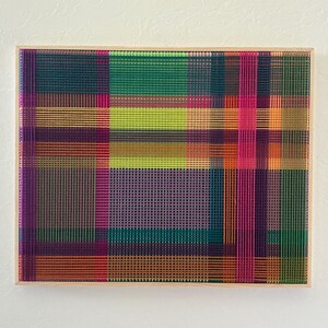 Könnte beinhalten: Abstrakte gewebte Kunstwerk in einem Holzrahmen. Das Stück zeigt ein gitterartiges Muster mit sich kreuzenden Linien in Grün-, Lila-, Orange-, Rosa- und Gelbtönen. Das Kunstwerk ist an einer weißen Wand befestigt.