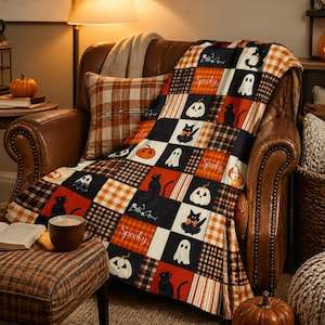 Op de afbeelding: Een patchwork deken met een herhalend patroon van zwarte katten, witte geesten, oranje pompoenen en het woord Spooky, afgewisseld met oranje, zwarte en witte ruitjes. De deken is gedrapeerd over een bruine leren fauteuil.