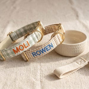 May include: Two pet collars featuring a gingham check pattern in sage green and beige, each equipped with gold-toned metal hardware. The sage collar is embroidered with the name MOLI in orange, while the beige collar features the name ROWEN in blue.
