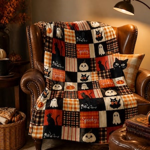 May include: A patchwork throw blanket featuring a repeating pattern of black cats, white ghosts, orange pumpkins, and skeletons, interspersed with orange, black, and white plaid squares. The word Spooky is printed in white script on several squares.