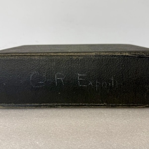 May include: A rectangular, black, leather-bound box with the handwritten text "G R Exp" on the side. The box has a slightly worn appearance, suggesting age or use. The background is a plain, light surface.