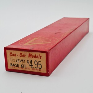 May include: A rectangular red cardboard box for a Con-Cor Models Tri-Level Auto Carrier base kit. The end label features the text Con-Cor Models, TRI-LEVEL BASE KIT, and the price $4.95. A gold triangular logo is printed on the top surface.