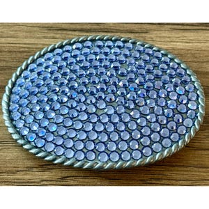 May include: An oval belt buckle featuring a dense arrangement of light blue, faceted circular crystals covering the entire surface. The buckle is framed by a metallic, twisted rope-style border that encircles the perimeter of the oval shape.