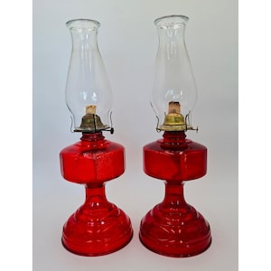 May include: Two matching oil lamps featuring vibrant red glass bases with a textured, faceted design. Each lamp includes a clear glass chimney, a brass-colored metal burner assembly, and a visible fabric wick centered within the burner.