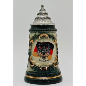 May include: A ceramic beer stein featuring a dark green base with gold trim and a metallic lid. The center displays a German flag with a black eagle emblem and the word Deutschland printed in black script. The body has ornate relief detailing.