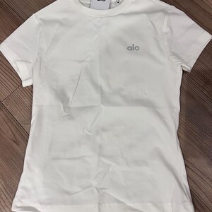 Puede incluir: Una camiseta blanca lisa de manga corta con cuello redondo. La prenda presenta la palabra alo impresa en una fuente gris peque&ntilde;a en la parte superior izquierda del pecho. Una etiqueta de marca rectangular blanca con el texto alo est&aacute; unida al cuello.