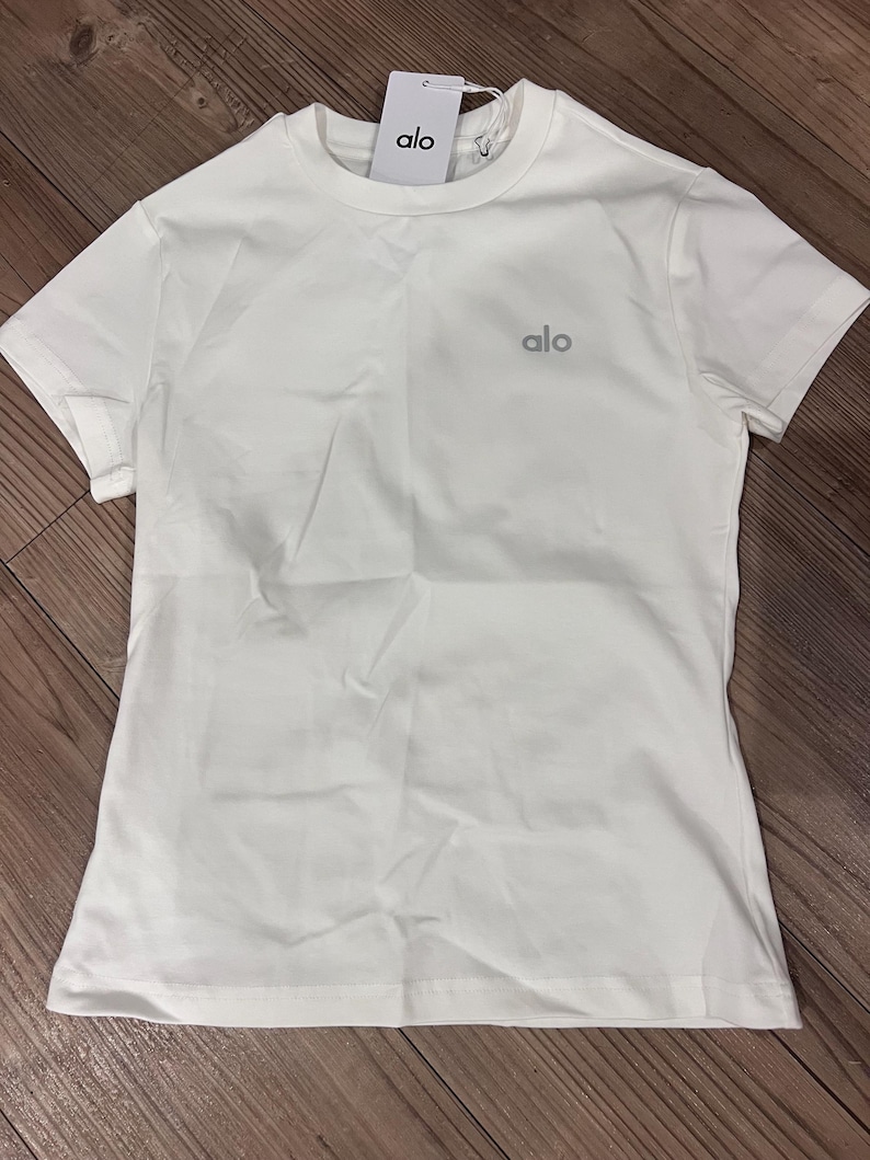 Puede incluir: Una camiseta blanca lisa de manga corta con cuello redondo. La prenda presenta la palabra alo impresa en una fuente gris peque&ntilde;a en la parte superior izquierda del pecho. Una etiqueta de marca rectangular blanca con el texto alo est&aacute; unida al cuello.