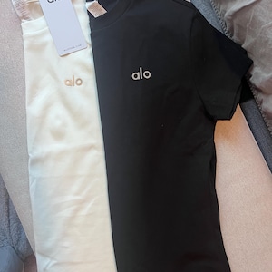 Puede incluir: Dos camisetas de manga corta y cuello redondo, una blanca y otra negra. Cada camiseta presenta la palabra alo bordada en una fuente peque&ntilde;a en la parte superior izquierda del pecho. La camiseta blanca incluye una etiqueta de papel con el texto alo y ALOYOGA.COM.