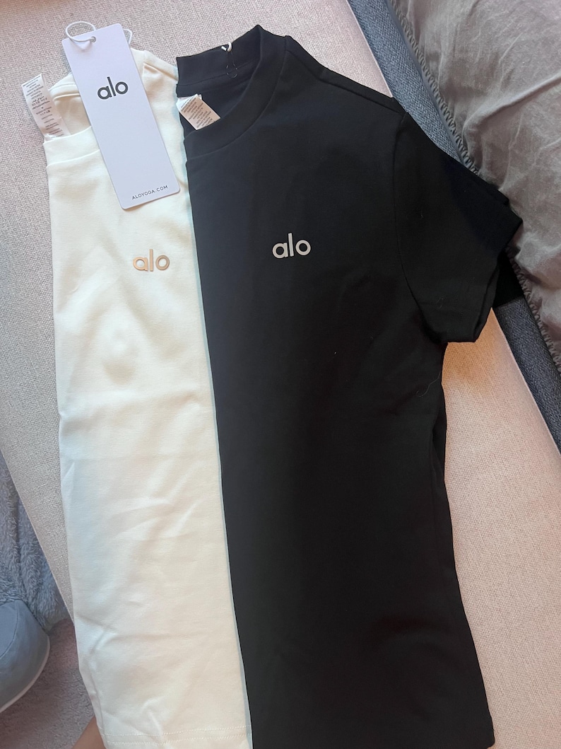 Puede incluir: Dos camisetas de manga corta y cuello redondo, una blanca y otra negra. Cada camiseta presenta la palabra alo bordada en una fuente peque&ntilde;a en la parte superior izquierda del pecho. La camiseta blanca incluye una etiqueta de papel con el texto alo y ALOYOGA.COM.