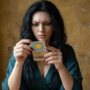 May include: A rectangular tarot card titled The Moon featuring a yellow sun, two dogs, and a lobster illustration. The card is held by hands with red nail polish, worn by an individual in a dark teal wrap top with a gold geometric necklace.