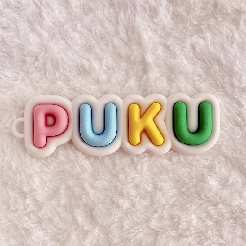 May include: A decorative charm featuring the word PUKU in raised, rounded letters. The letter P is pink, U is light blue, K is yellow, and the final U is green. Each letter is set against a white, cloud-like, scalloped border background.