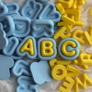 May include: A collection of small, three-dimensional alphabet letters in light blue and bright yellow. The letters are scattered, with the letters A, B, and C prominently displayed in the center, featuring yellow characters set against light blue frames.