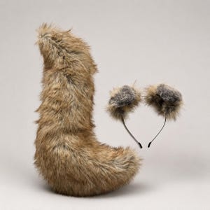 May include: A long, curved, faux fur tail in shades of brown and tan, paired with a thin black headband featuring two rounded, fluffy faux fur ears with dark gray centers. The set is designed for costume wear and features realistic textures.