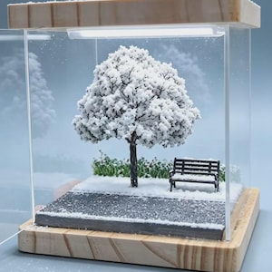 May include: A miniature diorama encased in a clear acrylic box with a light wood base. Inside, a dark-trunked tree with white foliage covered in snow stands next to a black park bench on a gray path, with green bushes dusted in white snow.