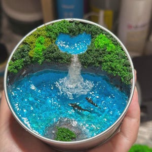 May include: A circular diorama in a metal tin featuring a miniature landscape with a heart-shaped blue pond at the top, a cascading waterfall made of clear resin, and a larger blue pool below containing two small black fish and a grey rock.