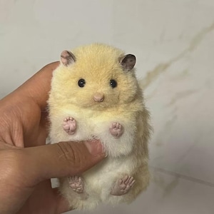Puede incluir: Una pequeña figura de hámster de peluche con pelaje color crema, ojos oscuros y pequeñas patas rosadas. El hámster tiene orejas redondeadas con puntas de color marrón oscuro y una nariz de color rosa claro. El pelaje se ve suave y denso.
