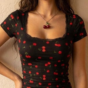 Puede incluir: Una camiseta corta negra de manga corta con un patrón repetitivo de cerezas rojas con tallos verdes. La prenda tiene un escote cuadrado adornado con encaje negro y se combina con un collar plateado con un colgante de cereza.