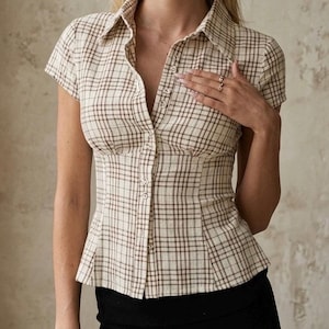 Peut inclure: Une chemise à manches courtes boutonnée avec un motif à carreaux beige et marron. La chemise présente un col structuré, un corsage ajusté avec des coutures verticales et une rangée de petits boutons sur le devant, jusqu'à l'ourlet.