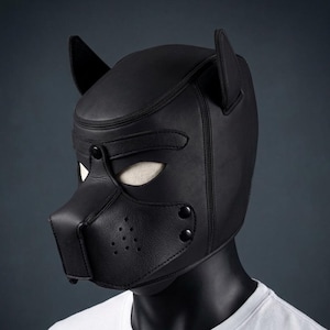 Pode incluir: Uma máscara de cachorro de couro preto com detalhes em forma de orelhas e um focinho. A máscara tem aberturas para os olhos, uma peça nasal com orifícios de ventilação e é fixada com rebites. A máscara é exibida em um manequim com uma camiseta branca.