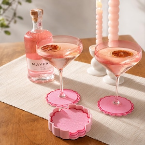 May include: Two pink scalloped coasters with a textured surface support two stemmed cocktail glasses filled with pink liquid and a dried citrus slice. A bottle of Mayfair Pink Gin and two tall pink candles are positioned behind the glasses.