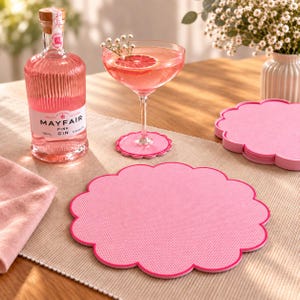 May include: A set of pink scalloped coasters with a textured surface, featuring a round shape with a decorative edge. The coasters are displayed alongside a bottle of Mayfair Pink Gin and a cocktail glass containing a pink drink with a grapefruit slice.