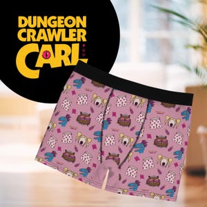 Puede incluir: Calzoncillos tipo bóxer con fondo rosa y un patrón repetitivo de personajes de dibujos animados, incluyendo un dinosaurio azul, un gato con gafas de corazón y un personaje rubio. La cintura es negra. El texto Dungeon Crawler Carl es visible.