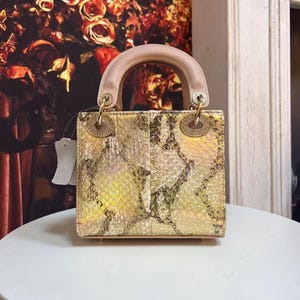 May include: A square-shaped handbag featuring a metallic gold and iridescent snakeskin-patterned finish. The bag has a structured beige leather top handle, gold-tone circular hardware attachments, and a small white tag attached to the side.
