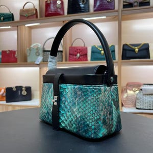 May include: A structured handbag featuring a black leather top flap and handle, paired with a textured teal and black snakeskin-patterned body. The bag includes a vertical black leather strap with a silver-toned metal clasp closure on the front.