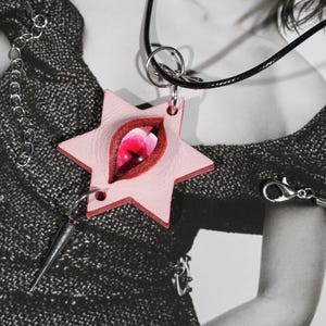 May include: A star-shaped pendant made of light pink material featuring a central, vertical, almond-shaped red glass eye. The pendant hangs from a black cord and includes a silver-toned metal spike charm attached to the bottom point of the star.