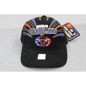 May include: A black baseball cap featuring embroidered text that reads NBA Champions. The design includes a circular Chicago Bulls logo with the years 91, 92, 93, 96, 97, and 98. Red, blue, and gold rays radiate from the center. A tag is attached to the side.