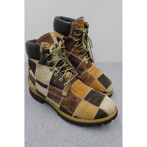 May include: A pair of ankle-high boots featuring a patchwork design of square leather panels in shades of tan, dark brown, and cream. The boots have a dark brown padded collar, yellow laces, metal eyelets, and a thick, dark brown rubber sole.
