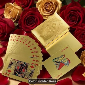 Puede incluir: Una baraja de cartas doradas con palos de diamantes rojos y una carta de comodín con una colorida ilustración de un bufón. Las cartas están desplegadas junto a una caja dorada a juego, todo sobre un conjunto de rosas rojas y doradas.