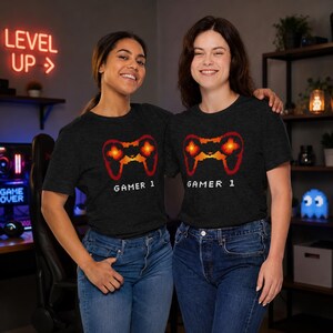 May include: A dark gray short-sleeve t-shirt featuring a pixelated orange and red video game controller graphic centered on the chest. Below the controller, the text "GAMER 1" is printed in a white, blocky, pixel-style font on the fabric.