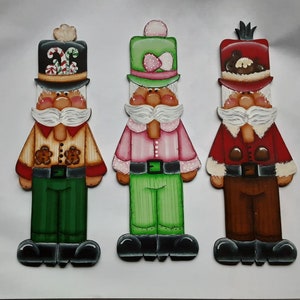 May include: Three wooden Christmas gnome ornaments, each with a different hat and outfit. The first gnome has a red and white candy cane hat, a tan shirt, and green pants. The second gnome has a green hat with a pink bow, a pink shirt, and green pants. The third gnome has a red hat with a brown fur trim, a red shirt, and brown pants.
