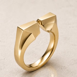 May include: A gold-toned open ring featuring a polished finish and two distinct, angular, geometric terminals that face each other with a small gap. The band is smooth and solid, showcasing sharp, faceted edges that create a structured, modern appearance.