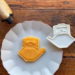 以下が含まれることがあります： ティーポットの形をした白いプラスチック製のクッキー型と、それと同じ形の黄色いクッキーが、縁が波打った白い皿の上に置かれています。クッキー型は、持ち手と注ぎ口があるティーポットの輪郭をはっきりと示しており、クッキーの形と一致しています。
