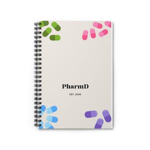 Op de afbeelding: Een spiraalgebonden notitieboek met een lichtbeige kaft met de zwarte tekst PharmD en EST. 2026 in het midden. De hoeken zijn versierd met illustraties van groene, roze, blauwe en paarse capsules gerangschikt in kleine groepjes.