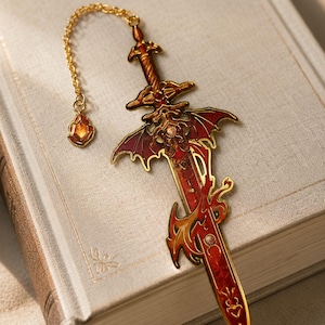 May include: A gold-toned metal bookmark shaped like a sword with red and orange enamel detailing. The sword features wing-like crossguards and a delicate gold chain attached to a small, faceted amber-colored charm at the end of the hilt.