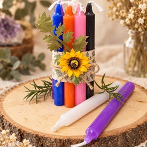 May include: A bundle of four tall taper candles in blue, orange, pink, and black, wrapped in a paper band with a yellow sunflower and green leaves. Two additional taper candles in white and purple lie horizontally in front of the bundle.