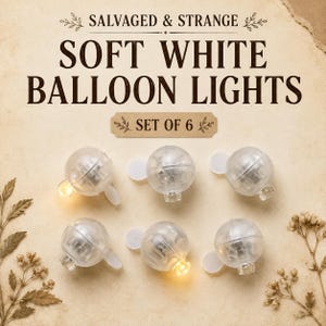 May include: A set of six clear, spherical balloon lights with internal LED components. The text SALVAGED & STRANGE SOFT WHITE BALLOON LIGHTS SET OF 6 is printed above the lights. Two of the lights are illuminated with a warm yellow glow.