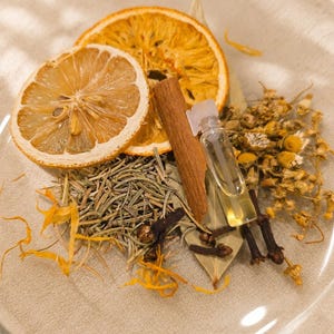 May include: A clear glass plate containing a collection of dried botanicals including two citrus slices, a cinnamon stick, a small glass vial of golden liquid, dried chamomile flowers, rosemary sprigs, whole cloves, and a single bay leaf.