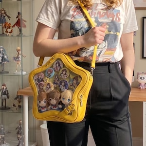 Puede incluir: Un bolso ita de color amarillo brillante con forma de estrella y una ventana frontal transparente. En el interior hay varios botones circulares con ilustraciones de personajes estilo anime y tres pequeñas muñecas de peluche en la base.